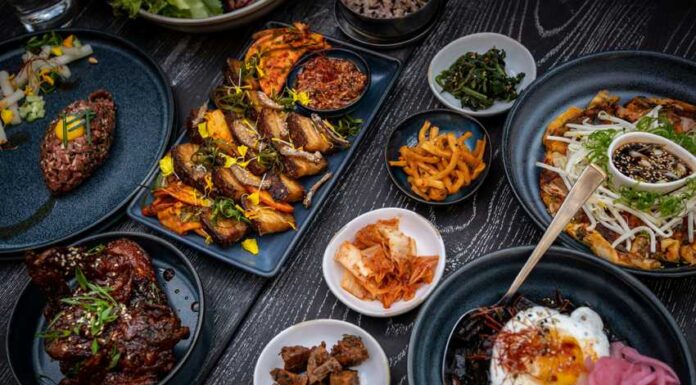 A variety of Korean dishes are arranged on a dark, textured table. The spread includes grilled meats, a large bibimbap bowl with a fried egg, various small side dishes (banchan) like kimchi and pickled vegetables, and a savory pancake. The plates are dark blue and black, contrasting with the vibrant colors of the food.