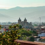 A scenic overlook of the historic cityscape of Oaxaca city, Mexico. The city is seen nestled in a valley surrounded by hazy mountains.