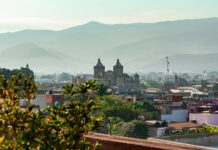 A scenic overlook of the historic cityscape of Oaxaca city, Mexico. The city is seen nestled in a valley surrounded by hazy mountains.