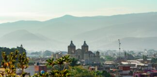 A scenic overlook of the historic cityscape of Oaxaca city, Mexico. The city is seen nestled in a valley surrounded by hazy mountains.