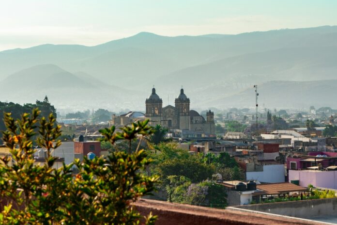 A scenic overlook of the historic cityscape of Oaxaca city, Mexico. The city is seen nestled in a valley surrounded by hazy mountains.