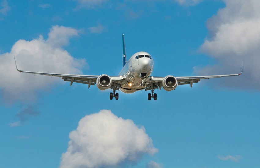 A silver colored unmarked passenger airplane in a blue sky with scattered puffy clouds. The plane's landing gear is down.