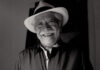 A black and white photo of a friendly elderly Mexican man with a wide, warm smile wearing a white fedora-style hat with a black band around it. He is wearing a dark colored button-down shirt. He has a gray mustache and gray hair.