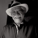 A black and white photo of a friendly elderly Mexican man with a wide, warm smile wearing a white fedora-style hat with a black band around it. He is wearing a dark colored button-down shirt. He has a gray mustache and gray hair.