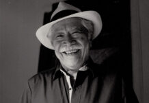 A black and white photo of a friendly elderly Mexican man with a wide, warm smile wearing a white fedora-style hat with a black band around it. He is wearing a dark colored button-down shirt. He has a gray mustache and gray hair.