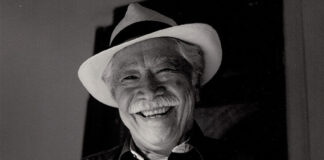 A black and white photo of a friendly elderly Mexican man with a wide, warm smile wearing a white fedora-style hat with a black band around it. He is wearing a dark colored button-down shirt. He has a gray mustache and gray hair.