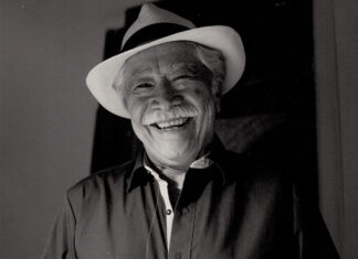A black and white photo of a friendly elderly Mexican man with a wide, warm smile wearing a white fedora-style hat with a black band around it. He is wearing a dark colored button-down shirt. He has a gray mustache and gray hair.