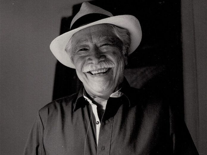 A black and white photo of a friendly elderly Mexican man with a wide, warm smile wearing a white fedora-style hat with a black band around it. He is wearing a dark colored button-down shirt. He has a gray mustache and gray hair.
