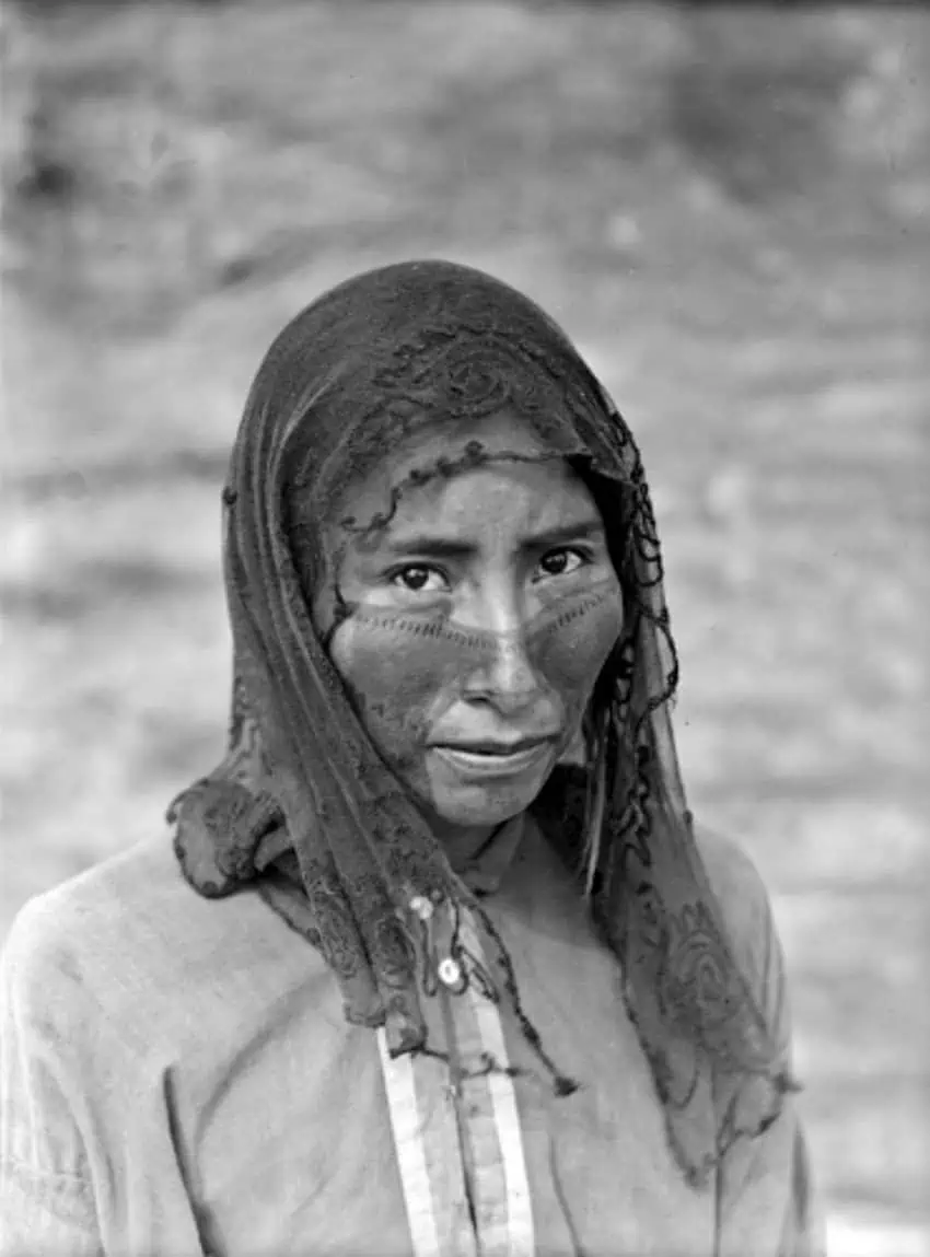 Black and white photo of a Raramuri woman wearing a traditional headscarf and button down traditional shirt. She looks straight into the camera. 
