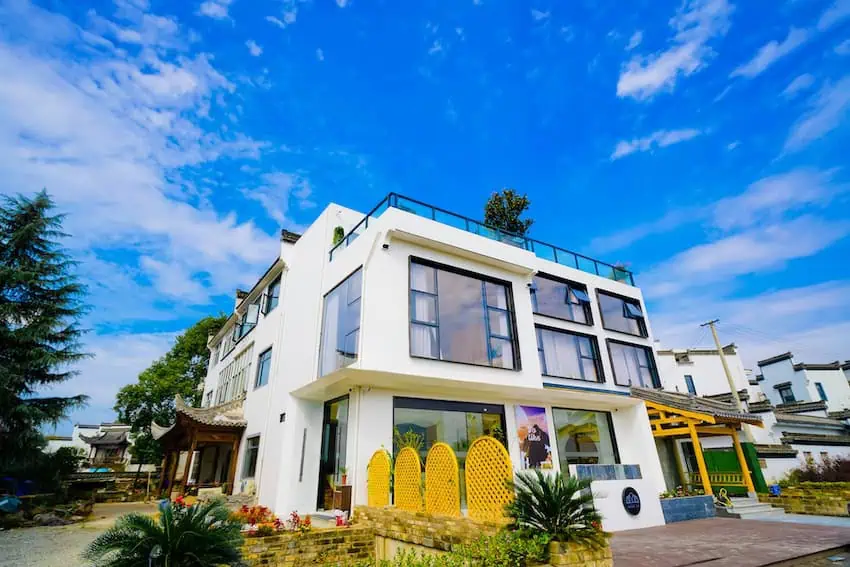 A modern white multi-story home with large windows, rooftop terrace, and colorful yellow decorative screens, surrounded by tropical plants under a bright blue sky.