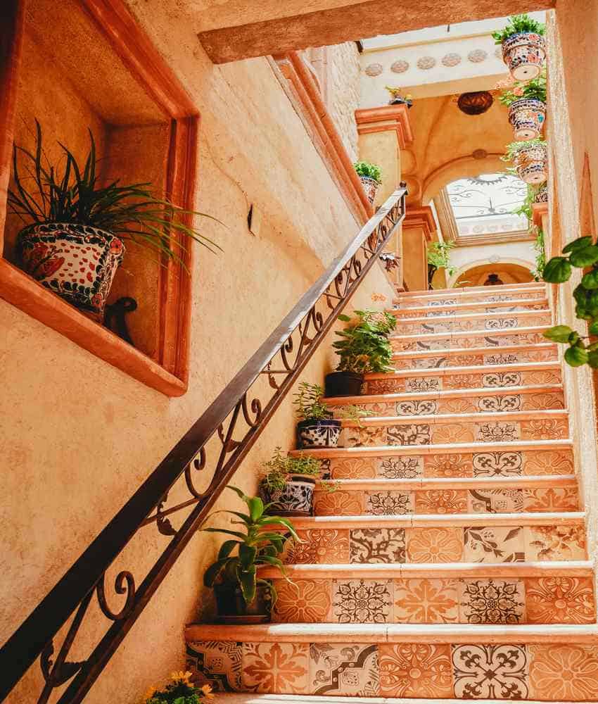 A colorful staircase in Mexico with each step decorated with a unique, patterned tile, leading up to a sunlit second-floor hallway with hanging potted plants. A wrought-iron handrail runs alongside the stairs, and a built-in wall niche holds a potted plant.