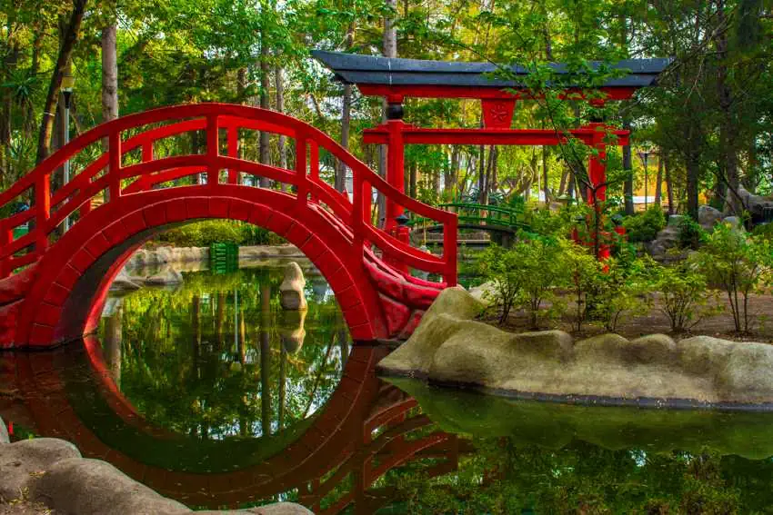 A vibrant, red arched footbridge and a traditional Japanese gate are reflected in the calm water of a pond in a Japanese garden in Mexico City.