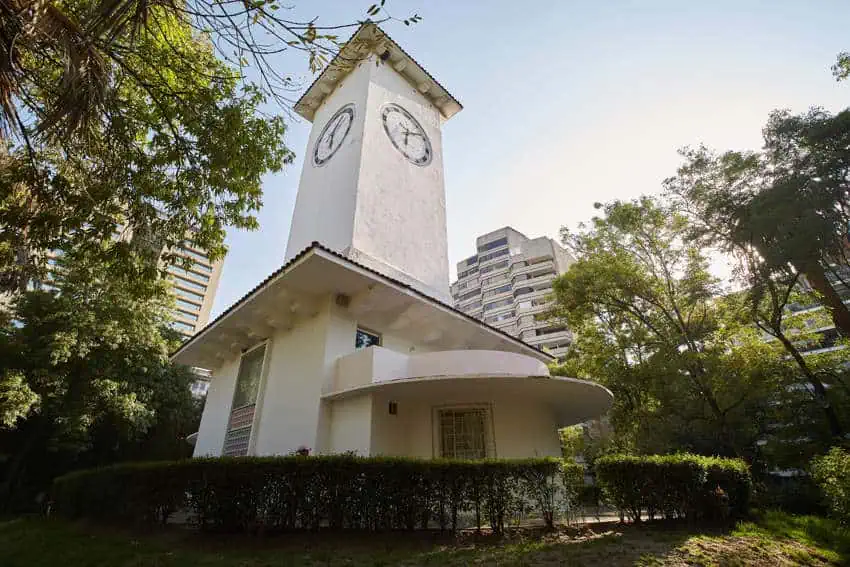 A white Art Deco clock tower viewed from a low angle on a sunny day in Parque Lincoln. In the background are surrounding trees and modern buildings of Mexico City.