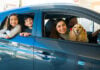 A smiling Mexican couple with two kids and a spaniel lean out of a small new car