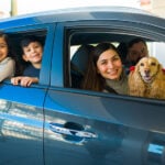 A smiling Mexican couple with two kids and a spaniel lean out of a small new car