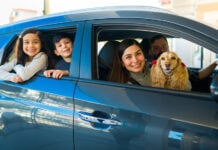 A smiling Mexican couple with two kids and a spaniel lean out of a small new car