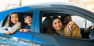 A smiling Mexican couple with two kids and a spaniel lean out of a small new car