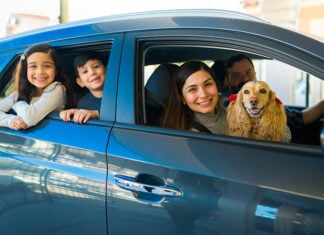 A smiling Mexican couple with two kids and a spaniel lean out of a small new car