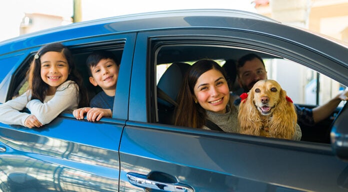 A smiling Mexican couple with two kids and a spaniel lean out of a small new car