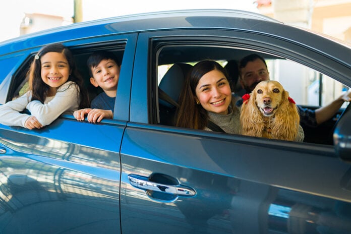 A smiling Mexican couple with two kids and a spaniel lean out of a small new car
