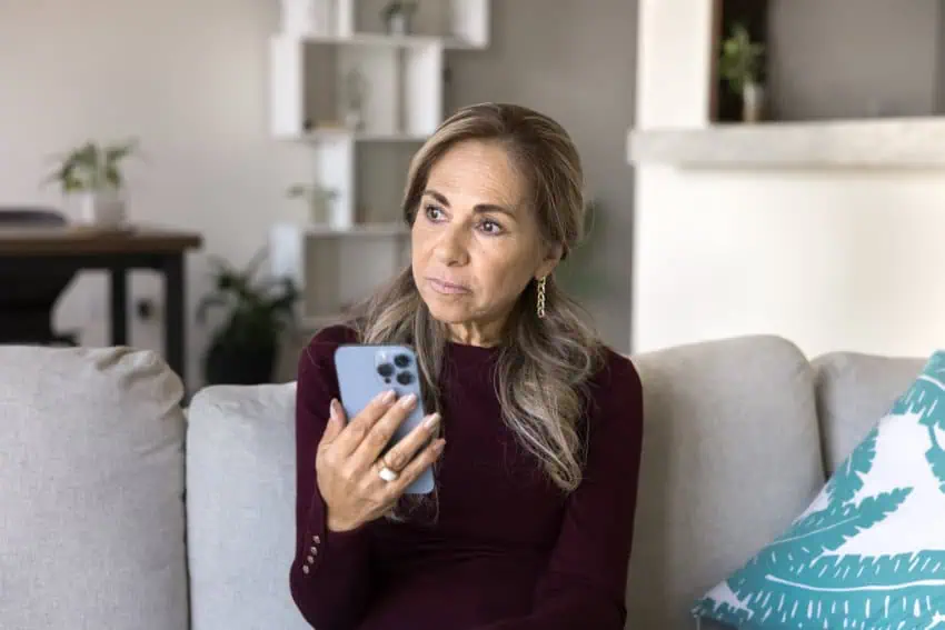Serious senior woman holds smartphone sits on sofa, looks away with thoughtful or concerned expression.