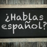 A small chalkboard showing the phrase "Hablas español?" in chalk. The chalkboard writing surface is black and it is framed by distressed unvarnished wood to look old, and is lying on a table or board made of similarly distressed wood.
