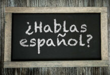 A small chalkboard showing the phrase "Hablas español?" in chalk. The chalkboard writing surface is black and it is framed by distressed unvarnished wood to look old, and is lying on a table or board made of similarly distressed wood.