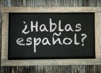 A small chalkboard showing the phrase "Hablas español?" in chalk. The chalkboard writing surface is black and it is framed by distressed unvarnished wood to look old, and is lying on a table or board made of similarly distressed wood.