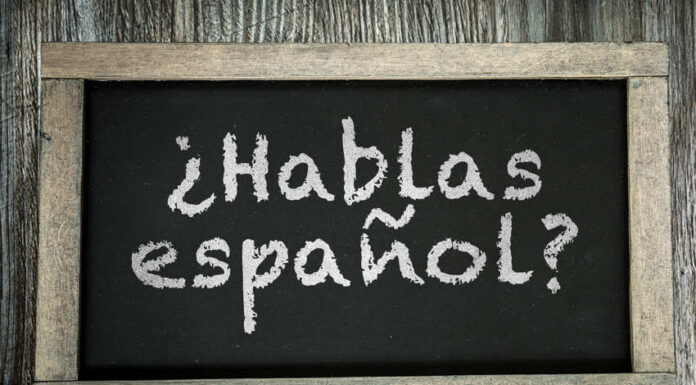 A small chalkboard showing the phrase "Hablas español?" in chalk. The chalkboard writing surface is black and it is framed by distressed unvarnished wood to look old, and is lying on a table or board made of similarly distressed wood.