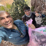 A selfie of two male hikers, one smiling at the camera and holding a mesh bag full of plastic bottles and other trash, with the second hiker behind him giving a thumbs up.