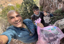 A selfie of two male hikers, one smiling at the camera and holding a mesh bag full of plastic bottles and other trash, with the second hiker behind him giving a thumbs up.