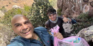 A selfie of two male hikers, one smiling at the camera and holding a mesh bag full of plastic bottles and other trash, with the second hiker behind him giving a thumbs up.
