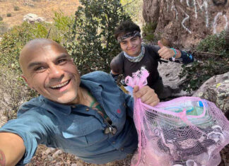 A selfie of two male hikers, one smiling at the camera and holding a mesh bag full of plastic bottles and other trash, with the second hiker behind him giving a thumbs up.
