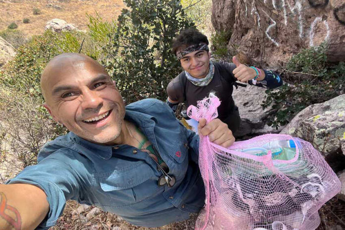 A selfie of two male hikers, one smiling at the camera and holding a mesh bag full of plastic bottles and other trash, with the second hiker behind him giving a thumbs up.