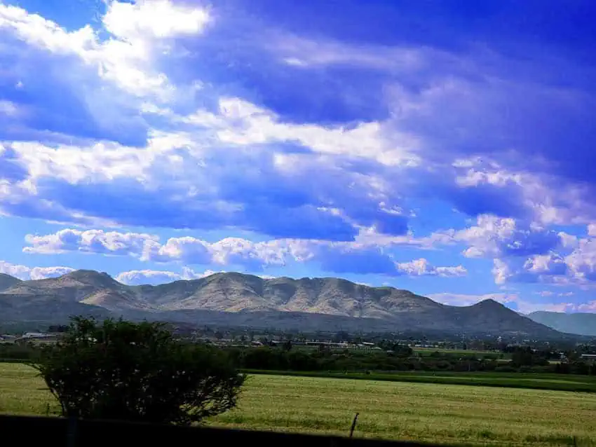 A panoramic view of the Cerro del Muerto mountain range in Aguascalientes, Mexico, with a verdant field in the foreground under a vibrant blue sky with scattered clouds.