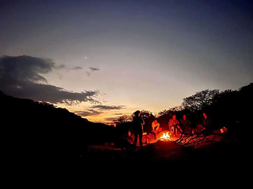 A group of friends sits around a campfire at dusk in a field in the state of Aguascalientes, Mexico. The setting sun illuminates the horizon with a warm, orange glow against a darkening sky.