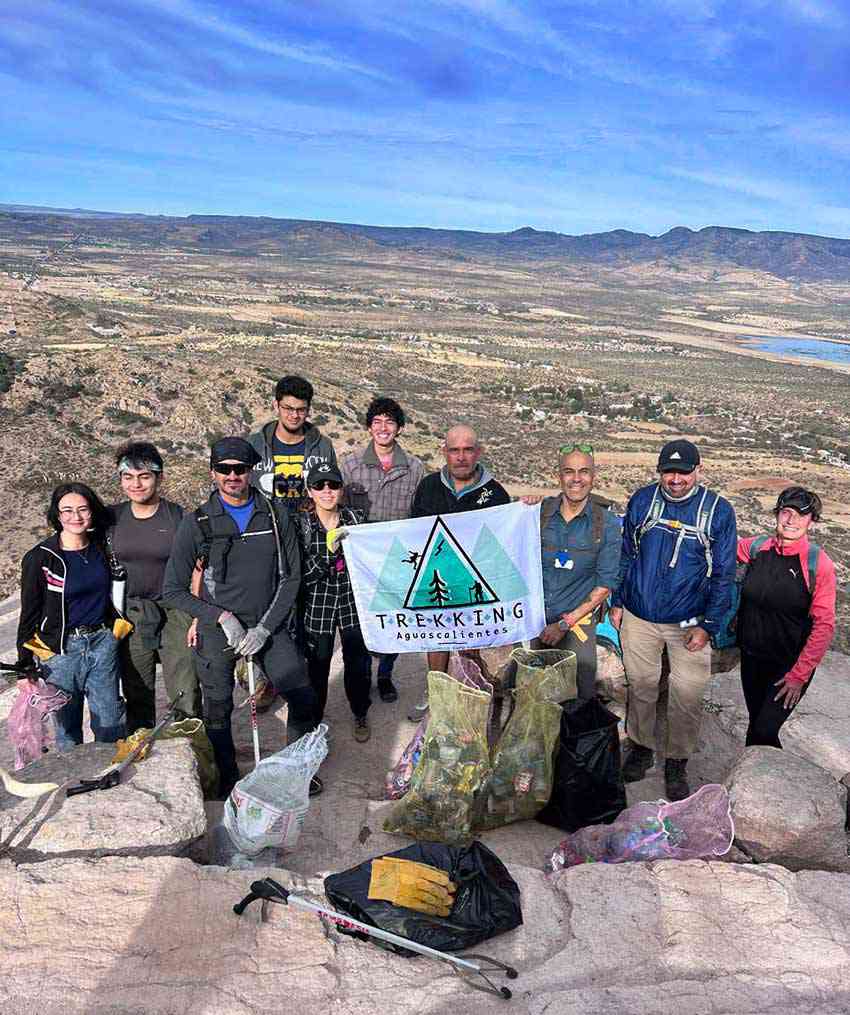 A group of people stands on a rocky summit, holding a flag that reads "Trekking Aguascalientes." They are surrounded by bags of trash they have collected, with a scenic, arid landscape and mountains visible behind them.