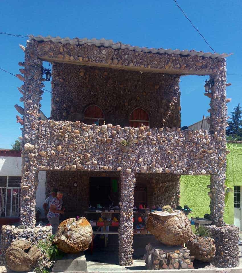 A unique two-story building in the town of Asientos, Aguascalientes, Mexico. The entire exterior is covered in small stones. The building has a small balcony and large, round boulders in front, with stuff for sale on the bottom floor, which is open-air.