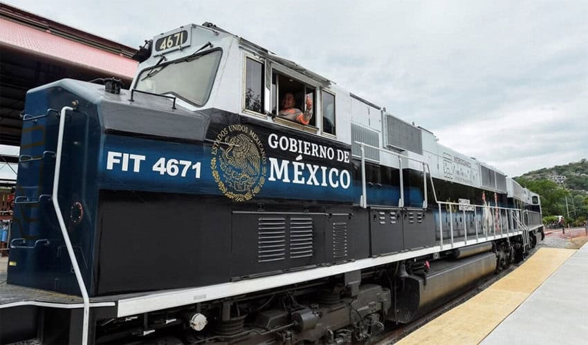 A modern blue and black locomotive of the Ferrocarril del Istmo de Tehuantepec is stopped at a station platform. The side of the train is prominently branded with the "Gobierno de México" logo, signifying its role in the Interoceanic Corridor infrastructure project.