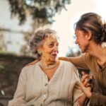 A caring younger woman provides support and companionship to an elegant elderly woman in a garden setting.