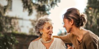 A caring younger woman provides support and companionship to an elegant elderly woman in a garden setting.