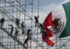 Workers install scaffolding in front of a giant Mexican flag
