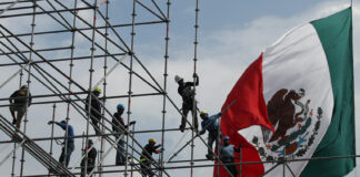 Workers install scaffolding in front of a giant Mexican flag
