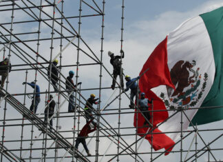 Workers install scaffolding in front of a giant Mexican flag