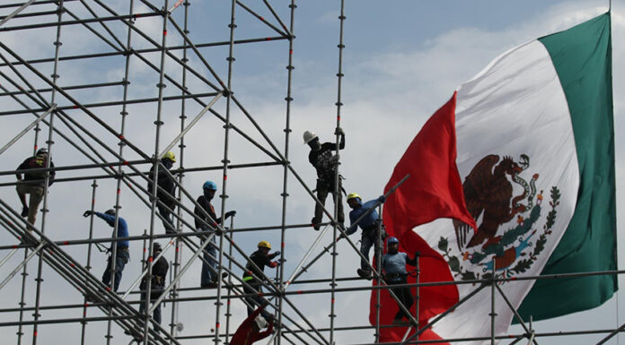 Workers install scaffolding in front of a giant Mexican flag
