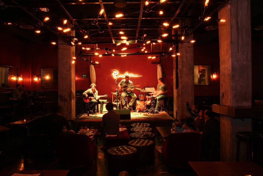 A jazz trio performs on stage under warm, glowing lights for an audience at Parker & Lenox, an intimate speakeasy-style jazz club in Mexico City.