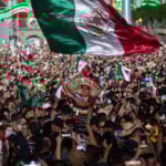 A man wearing a "Viva Mexico" sombrero wavves a giant Mexican flag in a packed crowd