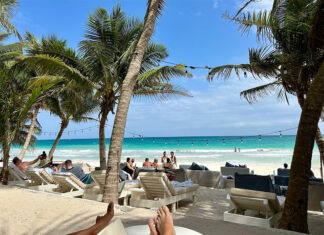 Lounge chairs and the occasional tourist fill a beach shaded by palm trees in Tulum