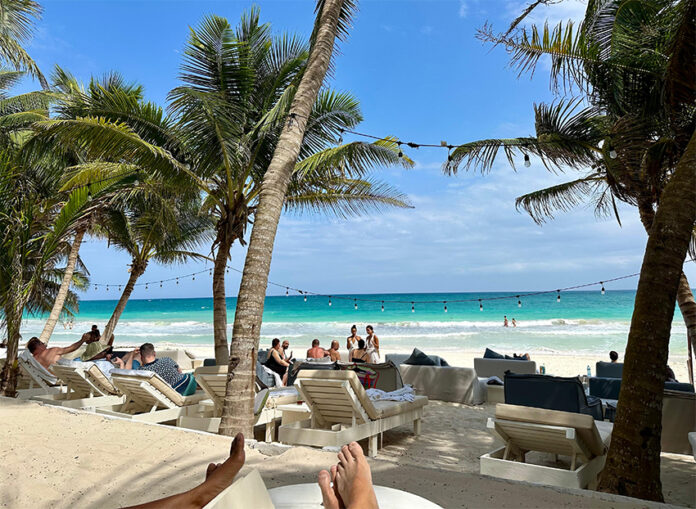 Lounge chairs and the occasional tourist fill a beach shaded by palm trees in Tulum