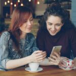 A woman with red hair is showing her phone to a woman with dark hair.